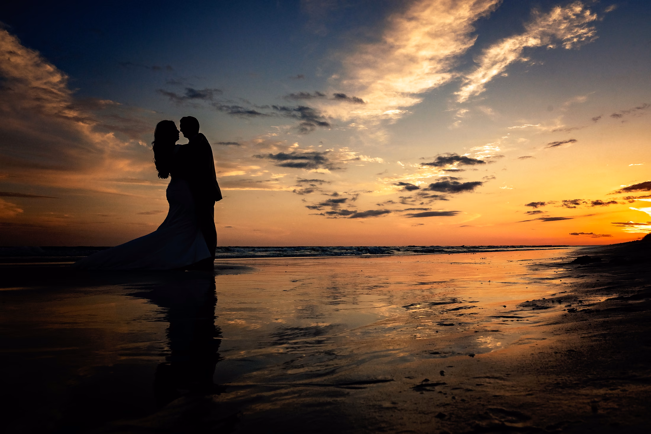 Couple kissing, silhouetted on the wet sand at sunset